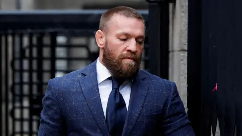 Reuters Conor McGregor, pictured from the chest up, wearing a navy tweed suit, a white shirt and a navy tie. He has tight brown hair and a brown beard. He is looking towards the ground. 