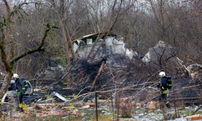 Emergency workers in protective gear examine debris at the site where a DHL cargo plane crashed into a house near Vilnius, Lithuania. The area is strewn with wreckage and fallen trees.