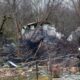 Emergency workers in protective gear examine debris at the site where a DHL cargo plane crashed into a house near Vilnius, Lithuania. The area is strewn with wreckage and fallen trees.