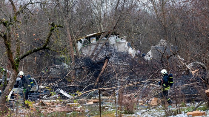 Emergency workers in protective gear examine debris at the site where a DHL cargo plane crashed into a house near Vilnius, Lithuania. The area is strewn with wreckage and fallen trees.