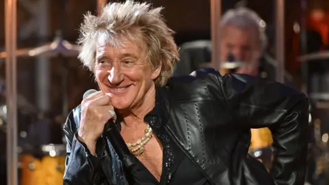 BBC Rod Stewart in a leather jacket and shirt unbuttoned halfway down his chest, holding a microphone and leaning to one side