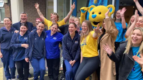 Tom Walker/BBC A group of nurses, some in uniform, and fundraising staff in blue and yellow Zoe's Place branded t-shirts, as well as a mascot in a yellow teddy-bear costume, smile and cheer 