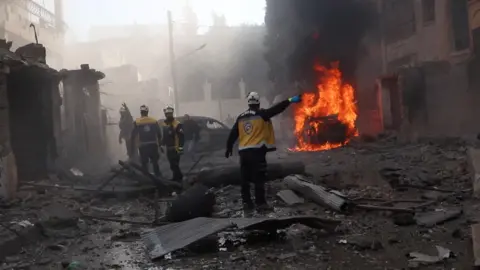 Reuters Rescue workers wearing white helmets and yellow jackets stand in a street covered in grey rubble, near a car engulfed in red flames. The air is full of smoke.