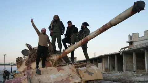 Getty Images Four men stand on top of an abandoned tank on the road to Aleppo. One man on the left has his hand raised in a v-for-victory style salute.