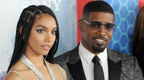 Getty Images Corinne Foxx with her long hair in braids worn down - she is wearing a silver halter-neck bra top. Jamie Foxx is to her left and is wearing a white shirt, black flecked jacket and black tie.