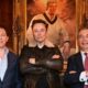 The three men stand below a portrait of a young Donald Trump wearing cricket gear