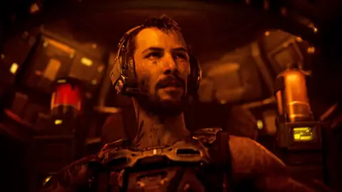 Prime Video A battle-scarred man sits inside the futuristic cockpit of a robot craft, illuminated in a dim orange light emanating from the controls. He wears a communications headset and a metal harness.