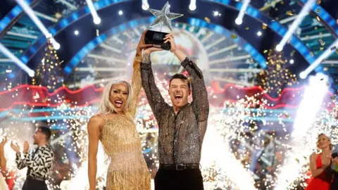 BBC Drag artist Tayce in gold dress lifts star-shaped trophy with dance partner Kai Widdrington on Strictly Come Dancing Christmas special as bright lights sparkle in the background