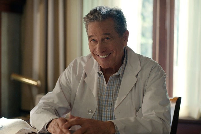 Tim Matheson as Doc, smiling ahead as he wears a doctor's lab coat and white shirt.