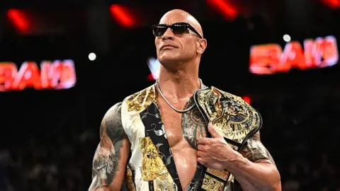 Getty Images The Rock standing in the middle of the ring, holding a golden title belt over his left shoulder, while wearing black sunglasses, a necklace chain and a white and gold top. Behind him is the 