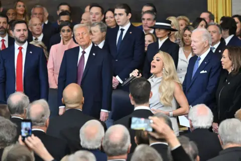 Getty Images Carrie Underwood, dressed in a silver dress, sings surrounded by people - including Donald Trump, Joe Biden and Kamala Harris, in suits