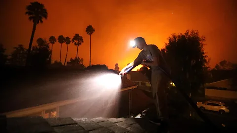 Getty Images A man douses the roof of his home with water from a hose while the sky glows orange with fire behind him (Credit: Getty Images)