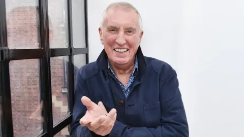 BBC Johnnie Walker smiling at the camera in BBC Old Broadcasting House. He is wearing a blue and white checked shirt and navy jacket.