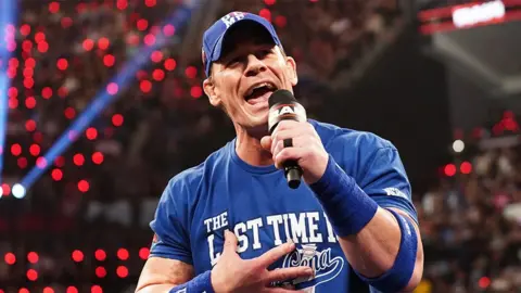 Getty Images John Cena, a man wearing a blue cap, t-shirt and sweat bands, speaking into a black microphone at WWE's Raw event in Los Angeles